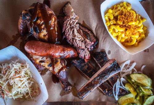 A platter of barbecue featuring smoked meats, sausage, macaroni and cheese, coleslaw, and pickles on a brown paper background.