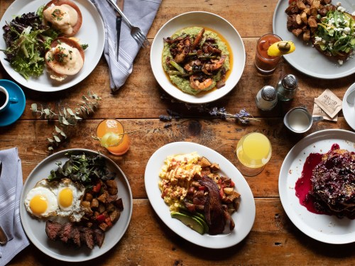 A spread of delicious brunch dishes on a wooden table, featuring eggs, meats, greens, and colorful drinks.