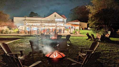 A cozy outdoor gathering at night, featuring a fire pit and a well-lit building with people enjoying the evening.