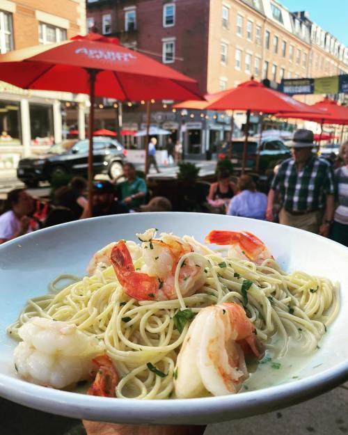 A bowl of shrimp pasta with a creamy sauce, held in front of a bustling outdoor restaurant with red umbrellas.