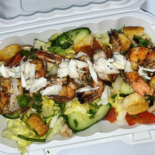 A takeout container filled with a salad topped with grilled shrimp, crab meat, croutons, cucumbers, and tomatoes.