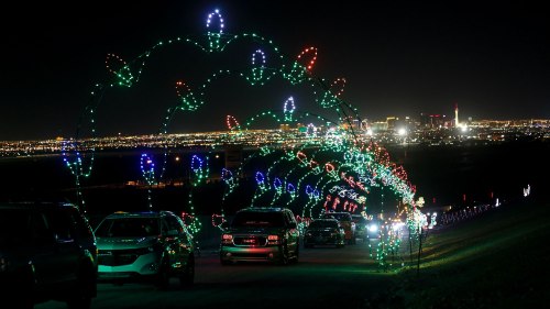 Colorful holiday lights form an arch over a road, with cars driving through and a city skyline in the background at night.