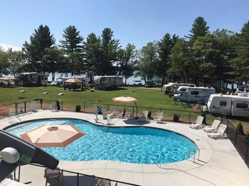 A sunny RV park with a swimming pool, surrounded by trees and parked RVs near a lake.