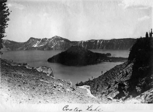Black and white photo of Crater Lake, featuring a calm lake surrounded by mountains and trees.
