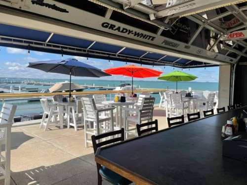 Outdoor dining area with white tables and colorful umbrellas overlooking a marina and blue water.