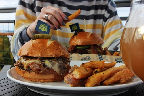 A close-up of two gourmet burgers with fries, one being lifted by a person, and a glass of beer in the foreground.