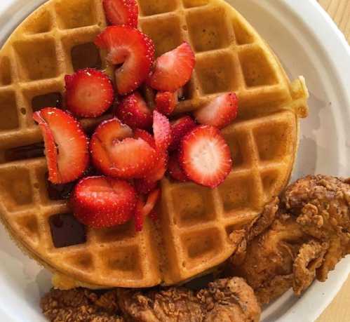 A golden waffle topped with fresh strawberries and syrup, served with crispy fried chicken on the side.