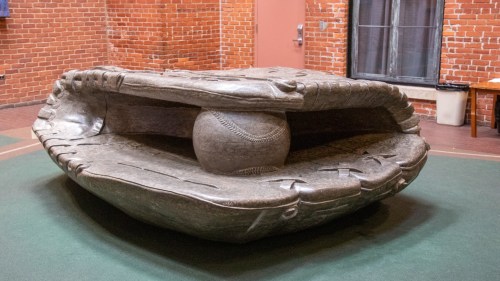 A large, sculptural baseball glove with a baseball nestled inside, set against a brick wall backdrop.
