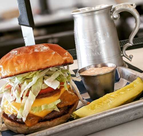 A juicy burger with lettuce, tomato, and cheese, served with a pickle and a small cup of sauce on a metal tray.