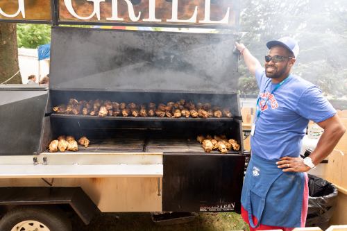 A man in a blue shirt and apron stands beside a large grill filled with cooking chicken, surrounded by smoke.