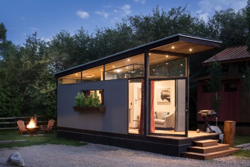A modern tiny house with large windows, surrounded by trees, featuring a fire pit and outdoor seating.
