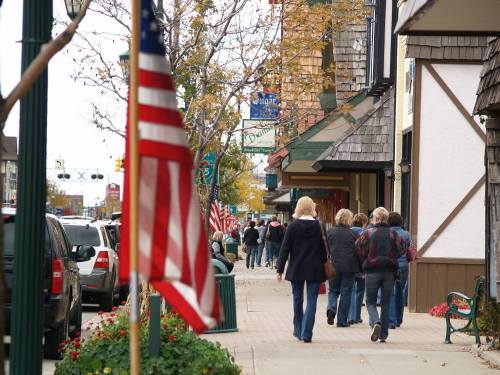 A busy street scene with people walking, shops lining the sidewalk, and American flags displayed.