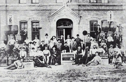A large group of people poses in front of a brick building, with barrels and signs, in a historical black-and-white photo.