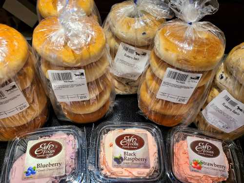 A display of packaged bagels stacked high, with containers of blueberry, black raspberry, and strawberry cream cheese nearby.