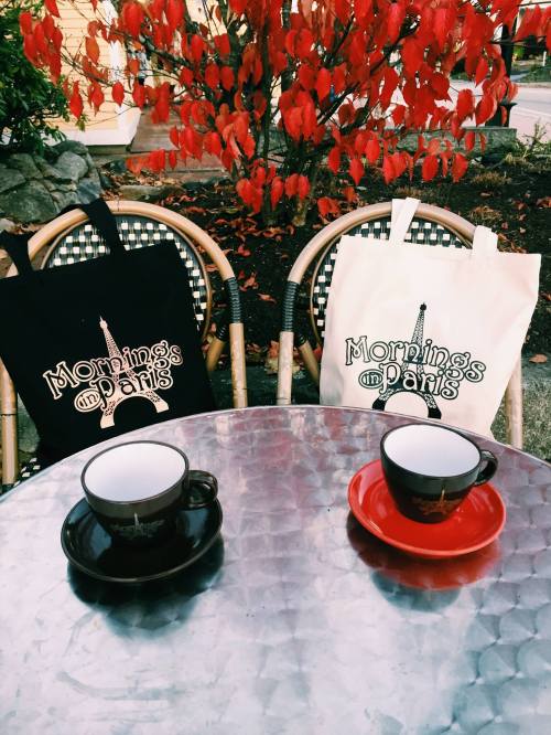 Two coffee cups on a metal table, with tote bags and vibrant red foliage in the background.