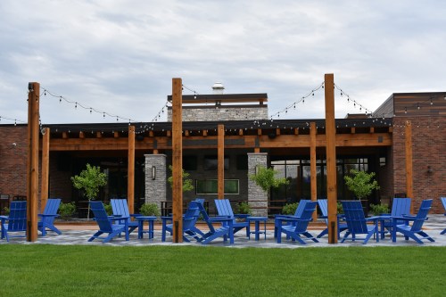 A patio with blue chairs and wooden posts, leading to a modern building with stone accents and string lights.