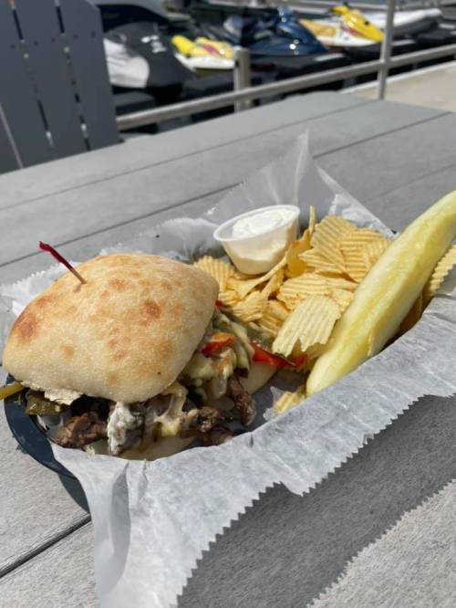 A sandwich with beef, peppers, and cheese, served with chips, a pickle, and a side of dipping sauce on a wooden table.