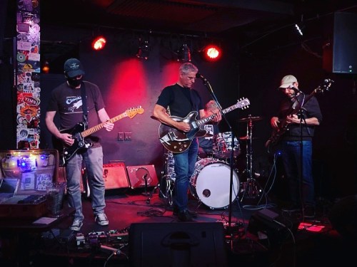 A band performs on stage with guitars and drums, surrounded by colorful lights and stickers on the walls.