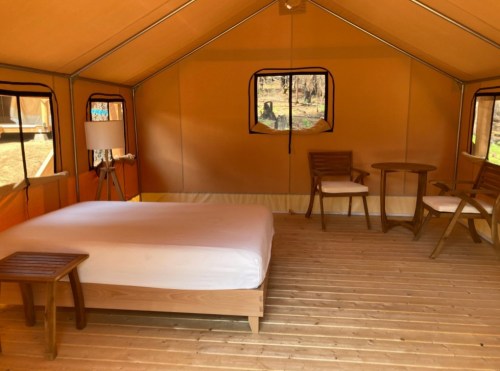 Interior of a glamping tent featuring a bed, wooden furniture, and large windows, set in a natural environment.