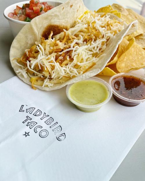 A taco filled with shredded cheese and toppings, served with chips, salsa, and a side of pico de gallo on a napkin.