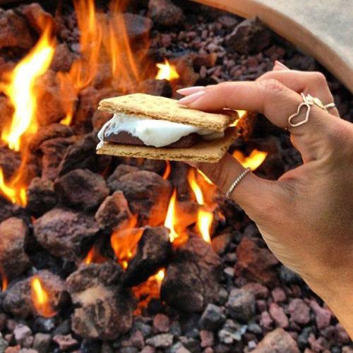 A hand holding a s'more with melted chocolate and marshmallow, above glowing embers and flames.