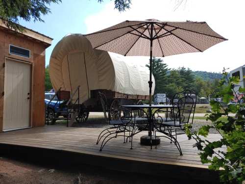 A wooden deck with a table and chairs under an umbrella, next to a covered wagon in a scenic outdoor setting.