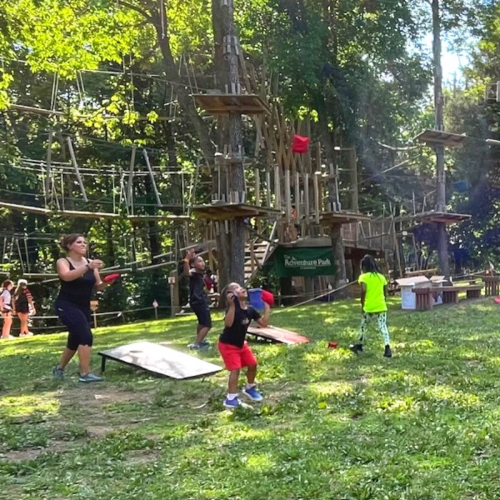 A group of children and adults play outside near a ropes course, surrounded by trees and green grass.