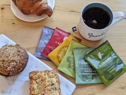 A cozy breakfast scene featuring a croissant, muffin, scone, coffee, and assorted tea packets on a wooden table.