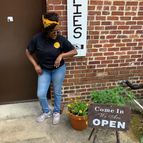 A woman in a black t-shirt and sunglasses stands by a "We Are Open" sign, with a potted plant nearby.
