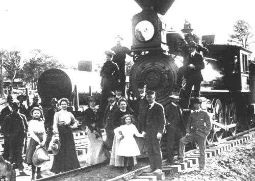 A historic black-and-white photo of a steam locomotive with a group of people, including children, posing on the tracks.