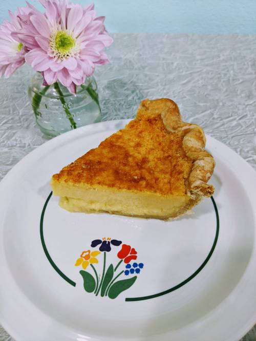 A slice of pie on a floral plate, accompanied by a small vase of pink flowers.