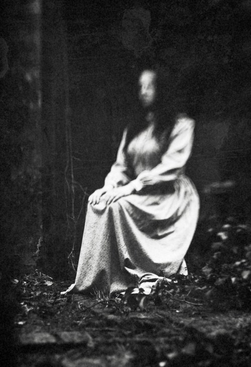 A blurred figure in a vintage dress sits on the ground, surrounded by dark foliage and shadows.