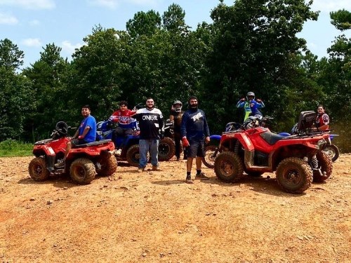A group of six people stands on a dirt hill with four ATVs, surrounded by trees and a blue sky.