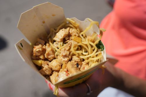 A hand holding a takeout box filled with chicken and noodles, garnished with vegetables.
