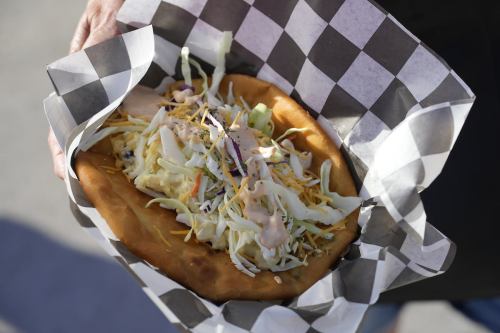 A hand holding a large, folded flatbread topped with shredded cabbage, cheese, and a creamy sauce, wrapped in checkered paper.