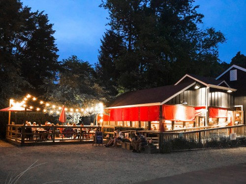 A cozy restaurant with red awnings and string lights, bustling with diners outdoors at dusk.