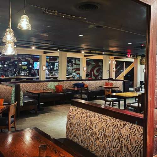 A cozy restaurant interior with wooden tables, patterned seating, and warm lighting, featuring a bar in the background.