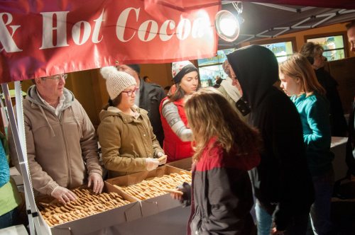 A festive market scene with people at a booth serving cookies and hot cocoa, surrounded by holiday decorations.