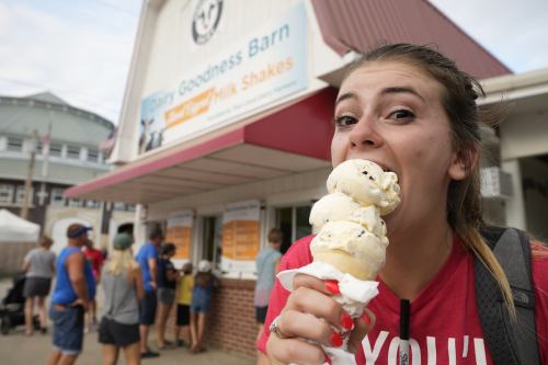 A young woman joyfully bites into a large ice cream cone outside a busy ice cream stand.