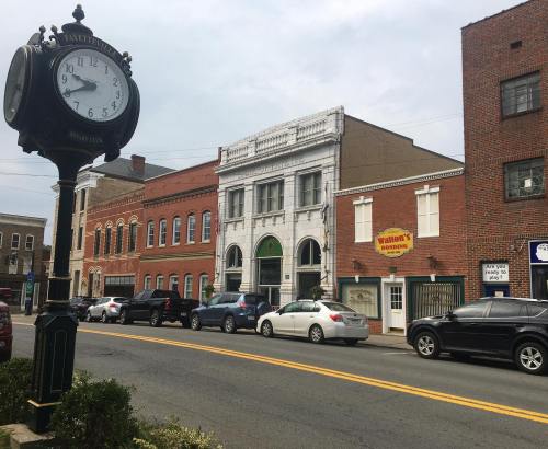 A street view featuring a vintage clock, brick buildings, and parked cars under a cloudy sky.