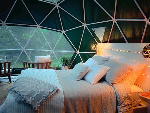 Cozy bedroom inside a geodesic dome, featuring a bed with pillows, soft lighting, and a view of nature through large windows.