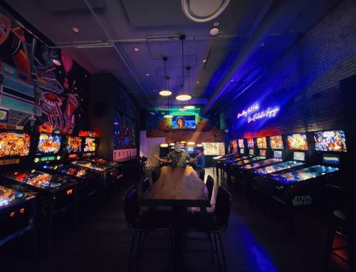 A vibrant arcade with pinball machines, neon lights, and a long table in a stylish, modern setting.