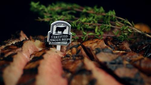 Close-up of grilled steak slices with a "Certified Angus Beef" sign and fresh thyme garnish.