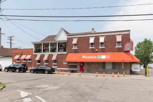 A brick restaurant with an orange awning and striped window shades, featuring a balcony with flowers and parked cars nearby.