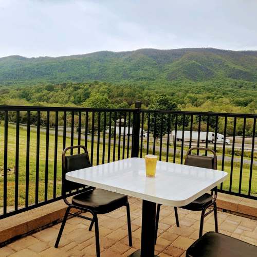 A table with a drink overlooks a green landscape and mountains in the background, with chairs nearby.