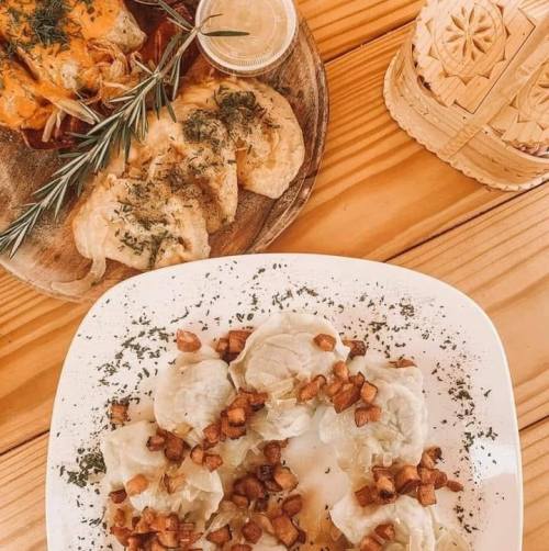 A wooden table with two plates of dumplings topped with herbs and crispy bits, alongside a small container of sauce.