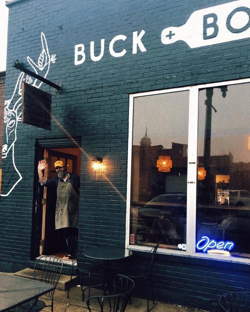 A person waves from the entrance of a cozy restaurant with a sign that reads "Buck + Boar" and an "Open" neon light.