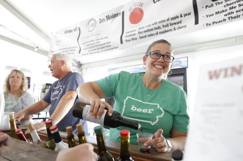 A woman in a green shirt pours a drink at a wine tasting event, with two others in the background.