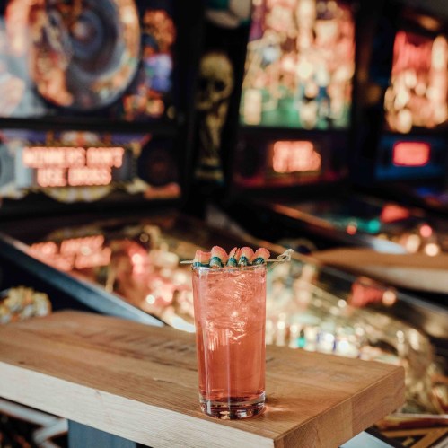 A vibrant cocktail with garnishes sits on a wooden table, with pinball machines and colorful lights in the background.