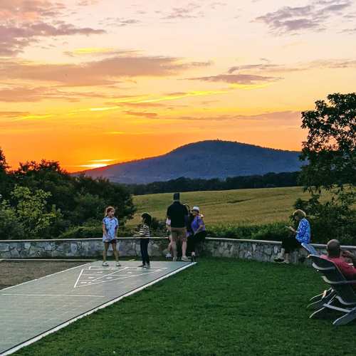 A sunset over rolling hills, with people enjoying the view from a grassy area near a stone wall.